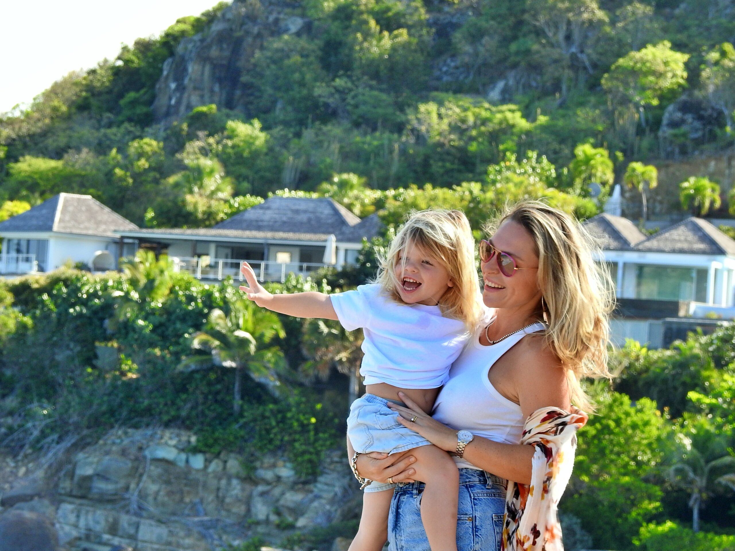 Photographie pleine de joie d’une femme tenant son enfant, avec un paysage tropical luxuriant en arrière-plan et une lumière naturelle éclatante.